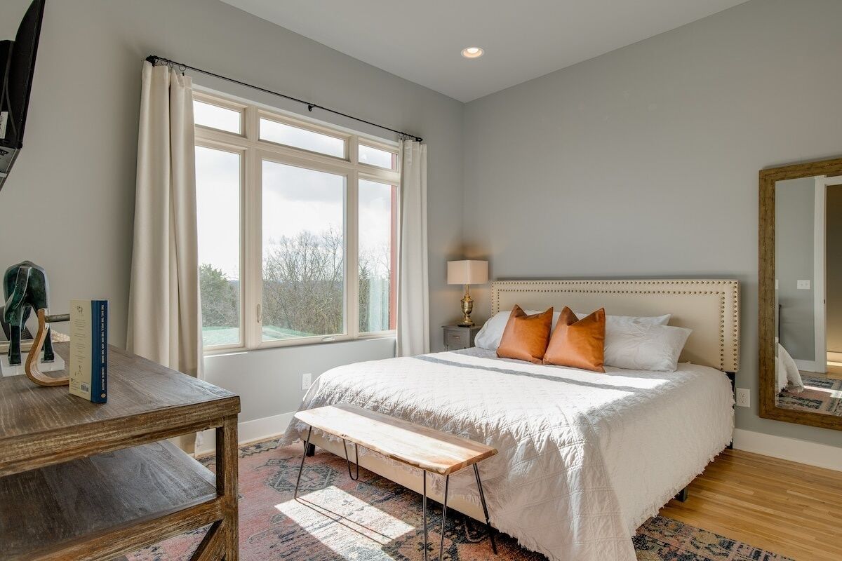 Une chambre avec un grand lit, une table de chevet, un banc, une fenêtre avec des rideaux, une lampe et un miroir au mur.