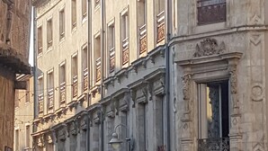 En byggnad med flera våningar, stora fönster och en balkong på höger sida. I förgrunden finns en gatlykta och en skylt med en röd cirkel och ett vitt streck.