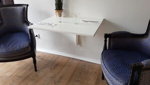 En matplats med ett vitt bord dukat för två personer, två stoppade stolar med ett blått mönster, en krukväxt på bordet och en vägg med en tydlig struktur i bakgrunden.