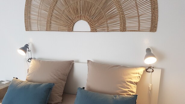 En säng med två beige kuddar och två blå kuddar, två sänglampor och en vävd väggdekoration ovanför.