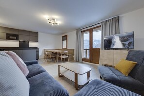 A modern living room with a sofa, a coffee table, and a TV. There is a dining area with a table and chairs. The room has a large window with curtains. - Ground floor, view lake, balcony, tv, ski locker, 56m², Tignes (Tignes)