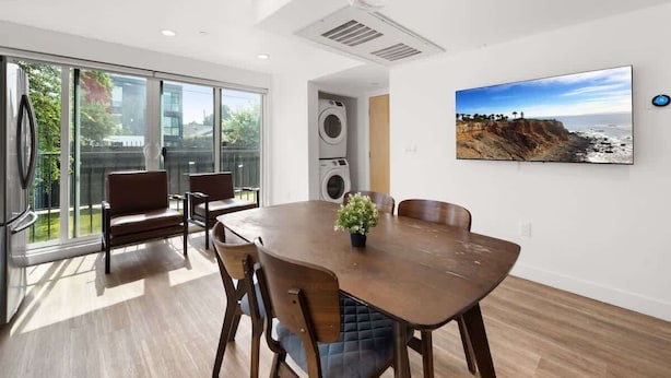 A modern dining area with a large table, chairs, and a potted plant. There is a large window with a view of greenery and a building. - Urban Retreat USC (Los Angeles)