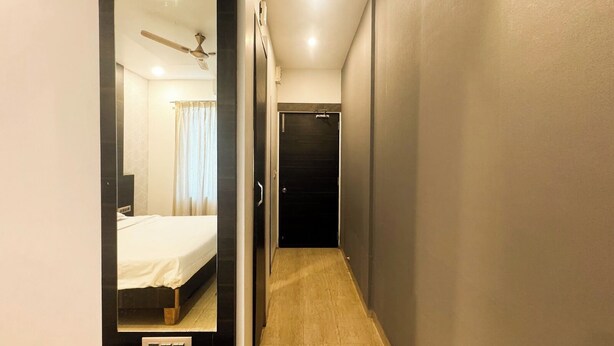 Un couloir étroit avec un mur recouvert de miroirs qui reflète une chambre à coucher. La chambre est équipée d’un lit, d’un ventilateur de plafond et d’une fenêtre avec des rideaux.