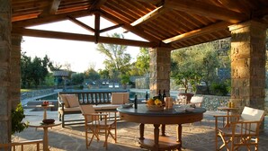 An outdoor patio with stone pillars, wooden furniture, and a pool in the background.