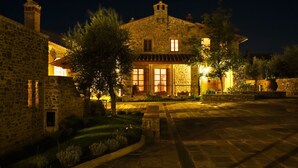 A stone building with multiple windows and a central entrance, surrounded by a paved courtyard and greenery.