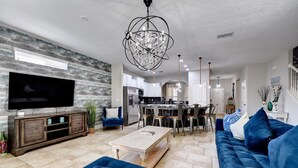 A modern living room with a large flat-screen TV, a blue sofa, a wooden coffee table, and a chandelier. - Luxurious Solterra Home w/ Pool, Spa & Themed Rooms Near Attractions (Davenport)