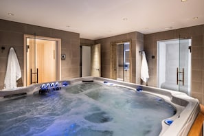 A modern spa room with a large hot tub, glass shower enclosures, and a wooden door leading to another room.