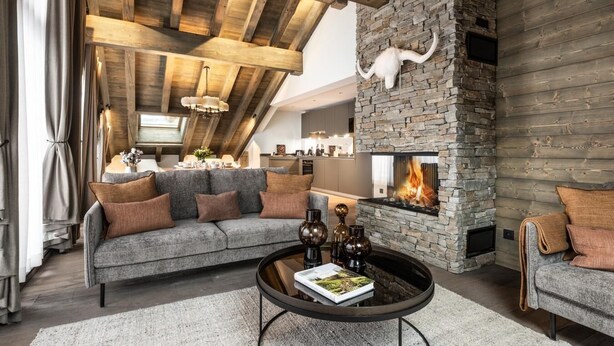 Un accogliente soggiorno con camino in pietra, soffitto in legno e un divano grigio con cuscini marroni.