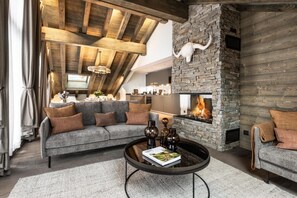 Uma sala de estar aconchegante com lareira de pedra, teto de madeira e um sofá cinza com almofadas marrons.