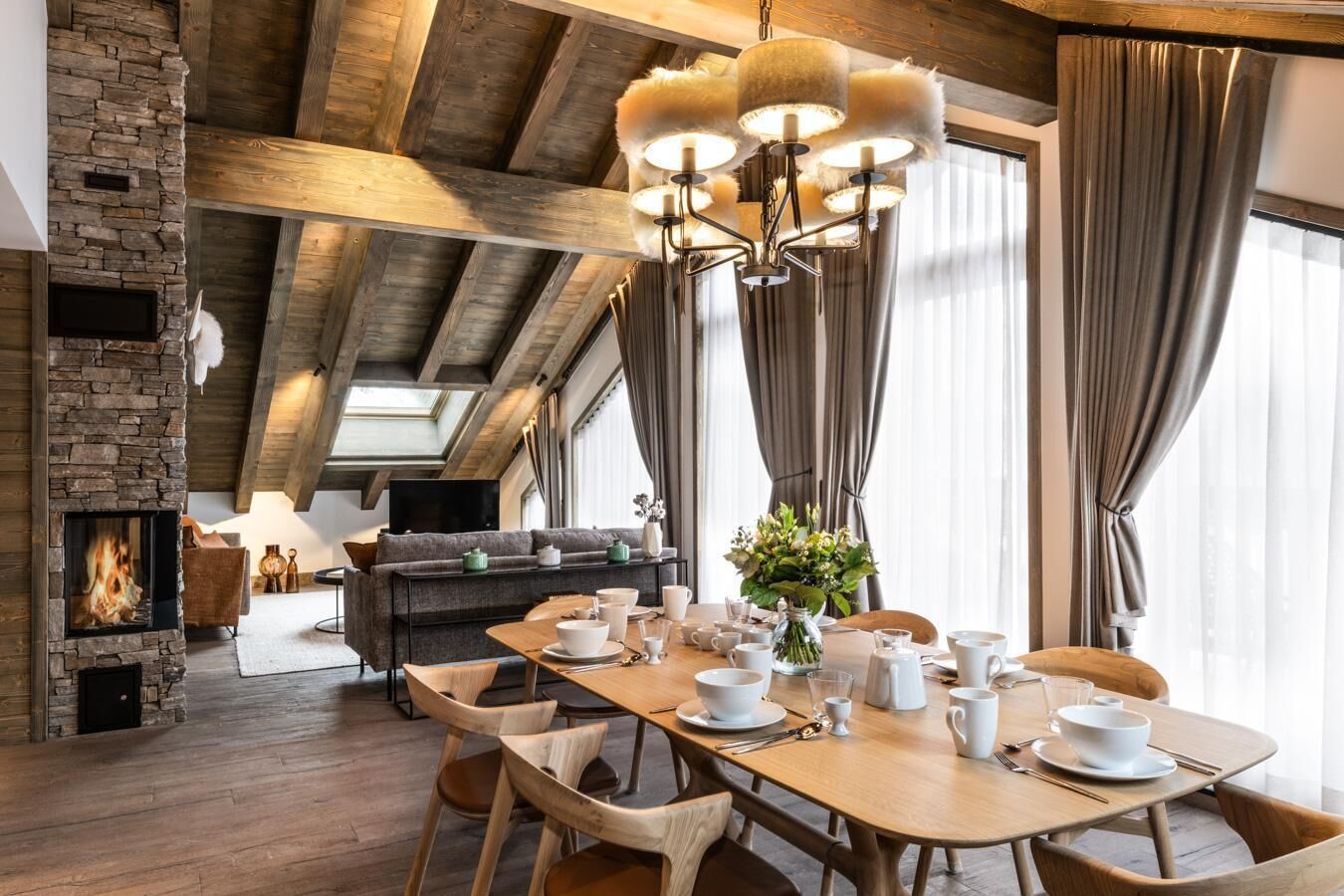 Een gezellige eethoek met een houten tafel gedekt voor vier personen, omringd door houten stoelen. Achtergrond toont een stenen open haard en een moderne bank.