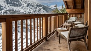 Una terraza de madera con vistas a montañas nevadas, sillas de mimbre y una pequeña mesa.