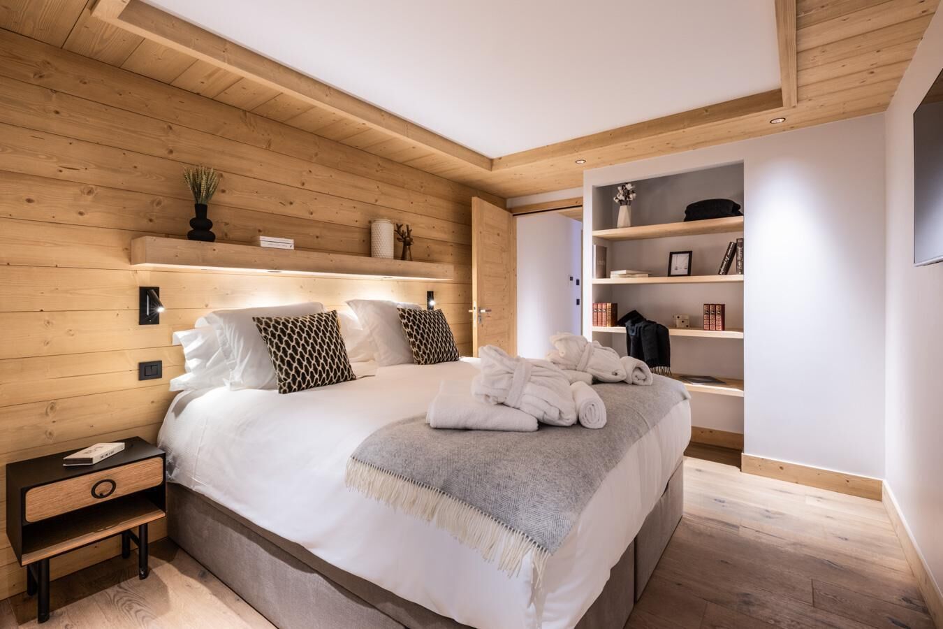 A modern bedroom with a wooden ceiling, a large bed with white bedding, a bedside table, and a built-in bookshelf.