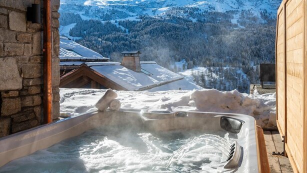 Un jacuzzi con vista a una montaña nevada, paredes de piedra y construcciones de madera.