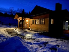 A wooden cabin with a covered porch and a snow-covered driveway at night.