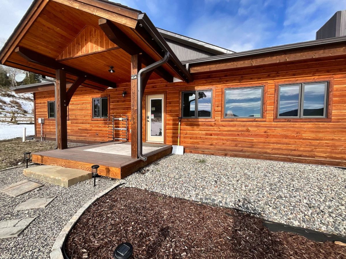A modern log cabin with a covered porch, large windows, and a gravel driveway.