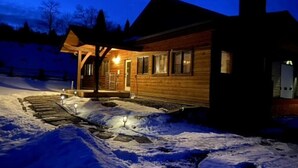 A wooden cabin with a covered porch and a snow-covered driveway at night. - NEW! LAST MINUTE SPECIAL! Spacious Modern Chalet, Close To Everything Big Sky, MT! (Big Sky)