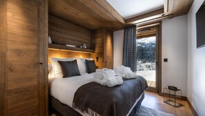 A modern bedroom with a wooden headboard, a large bed with white linens, a dark throw blanket, and a view of a snowy landscape through a large window. - Wifi, 150m from ski slopes, Ground floor, sauna, hammam, terrace, balcony, tv, 182m², Méribel (Méribel - Les Allues)