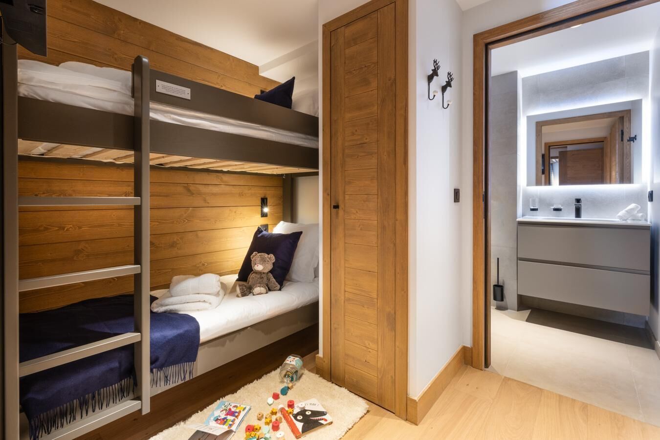 A dorm-style room with bunk beds, a small bedside table with a teddy bear, and a bathroom with a mirror and sink.