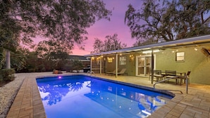 A swimming pool with a brick deck, surrounded by a covered patio area with chairs and a table, and trees in the background. - HUGE HEATED SALT POOL! NEAR DOWNTOWN! TV IN EVERY BEDROOM! (Sarasota)