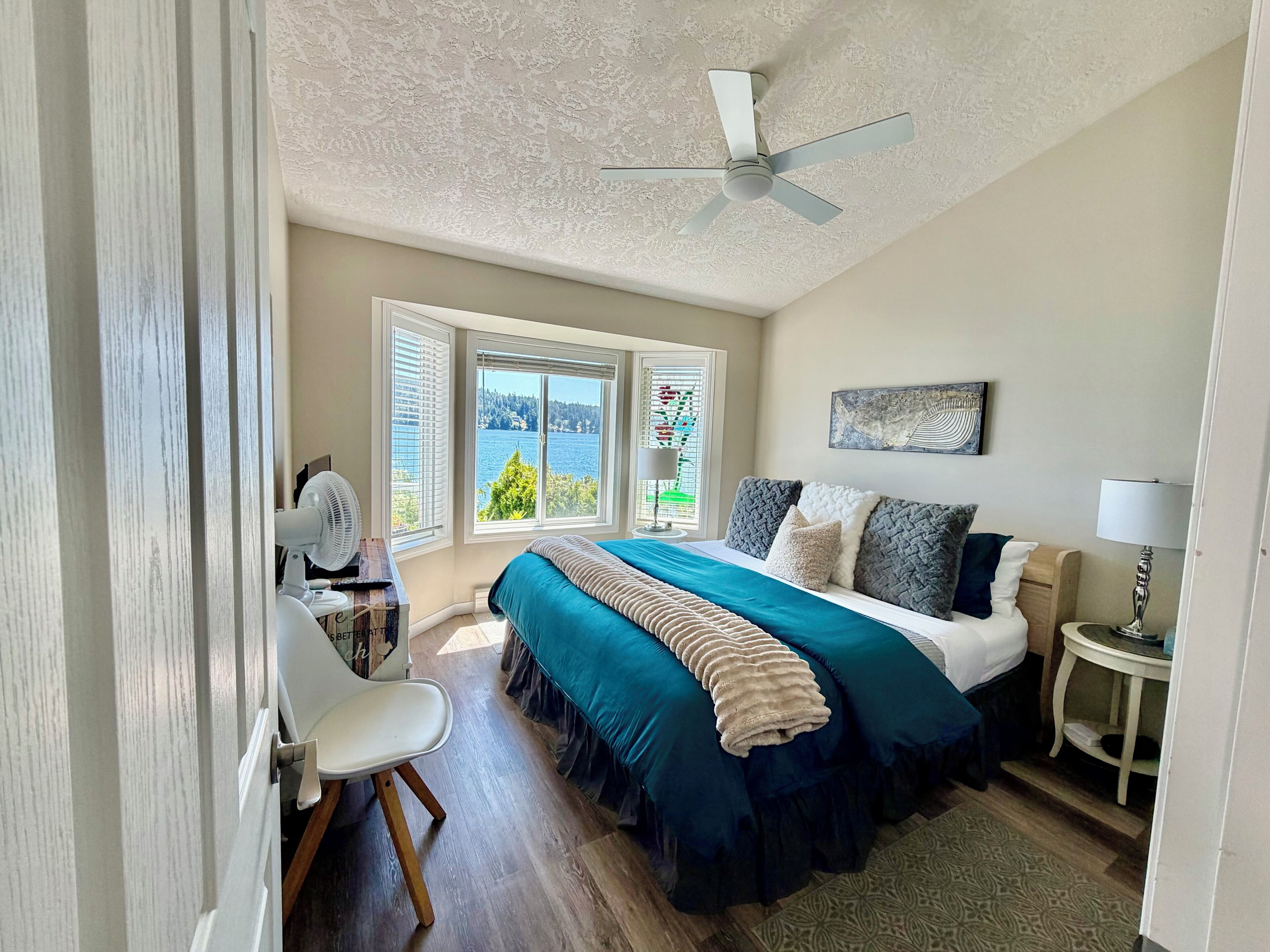 A bedroom with a bed, a ceiling fan, a desk with a chair, a lamp, and a window with a view.
