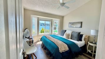 A bedroom with a bed, a ceiling fan, a desk with a chair, a lamp, and a window with a view.