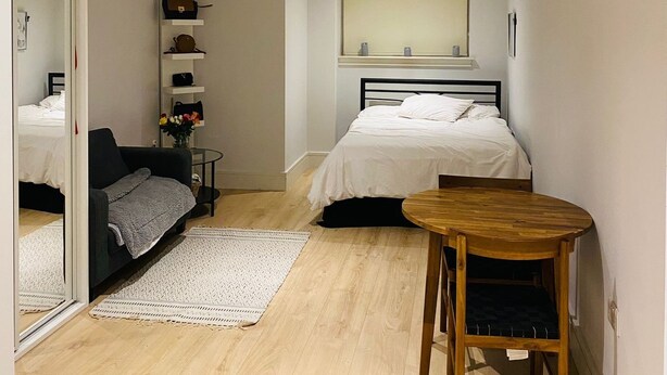 Et soveværelse med seng, et lille bord, en sofa, et spejl og en hylde med tasker.