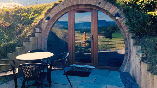 An outdoor patio with a round table and chairs, a wooden door leading to a view of a green landscape and mountains. - Honeybee Home -  a hobbit hut that sleeps 4 guests  in 2 bedrooms (Llansantffraid)
