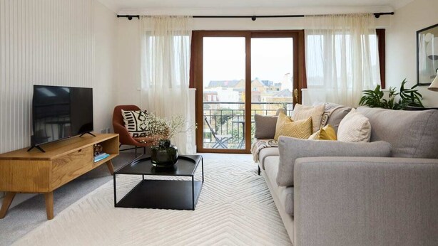 A modern living room with a sofa, a coffee table, a TV, and a balcony with curtains. - The Isleworth Place - Stunning 1bdr Flat With Balcony (Isleworth)