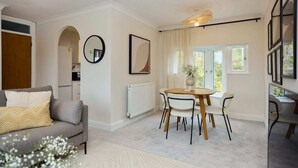 A modern living room with a grey sofa, a dining table with four chairs, a radiator, and a window with curtains. - The Isleworth Place - Stunning 1bdr Flat With Balcony (Isleworth)