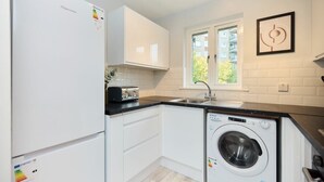 A compact kitchen with white cabinets, a built-in washing machine, a toaster, and a window with a view of buildings. - The Isleworth Place - Stunning 1bdr Flat With Balcony (Isleworth)