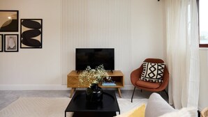 A modern living room with a flat-screen TV on a wooden stand, a black coffee table with a vase of flowers, a brown armchair with patterned cushions, and a large window with sheer curtains. - The Isleworth Place - Stunning 1bdr Flat With Balcony (Isleworth)