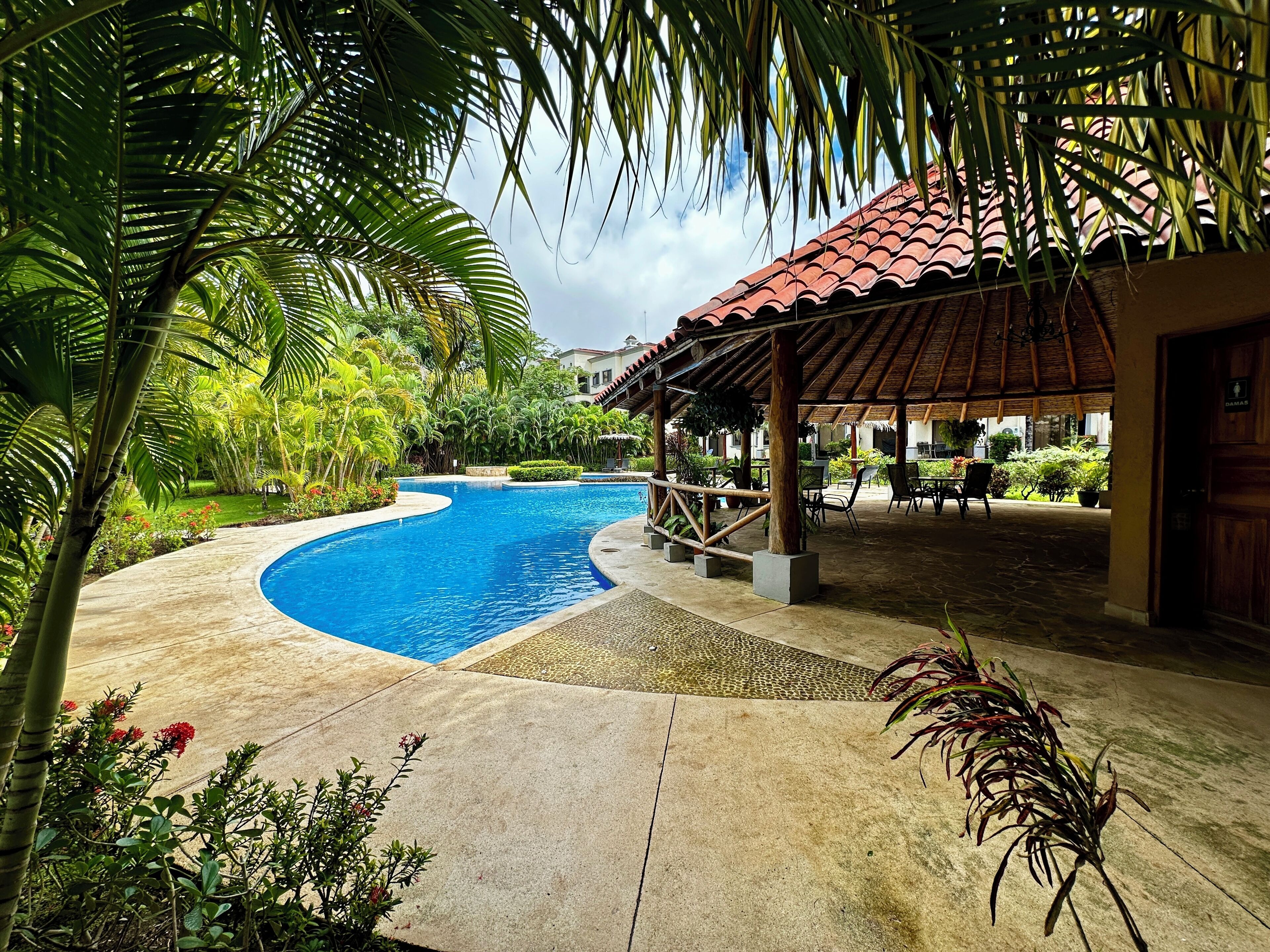 A swimming pool with a curved edge, surrounded by lush greenery and a covered seating area with chairs and tables.