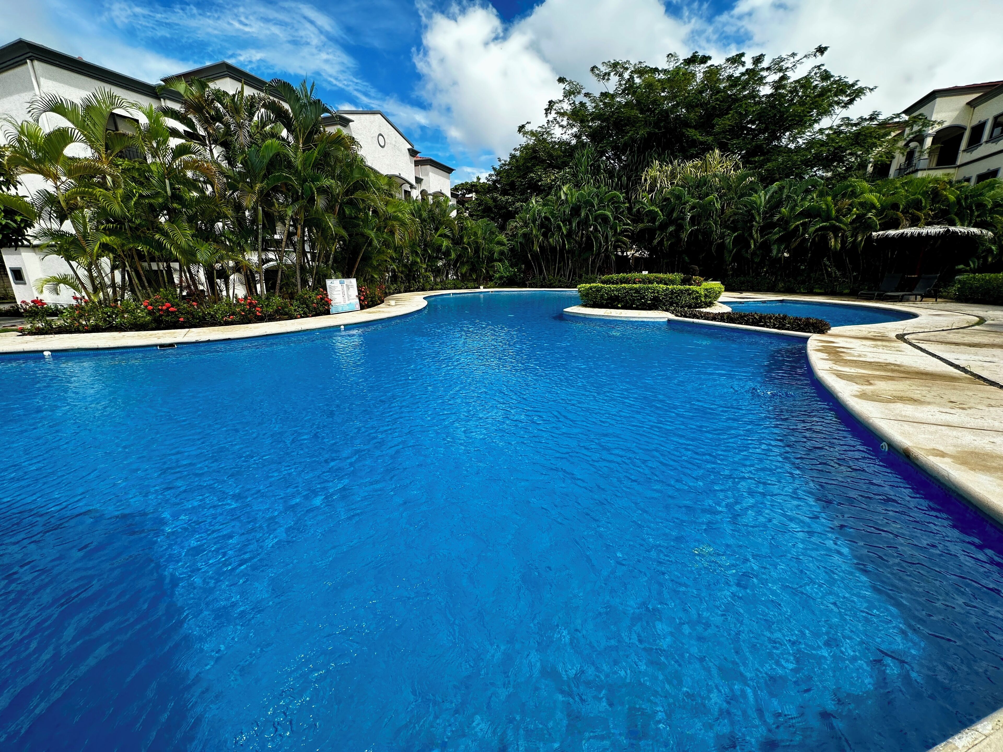 A large, clear blue swimming pool surrounded by lush greenery and white buildings.