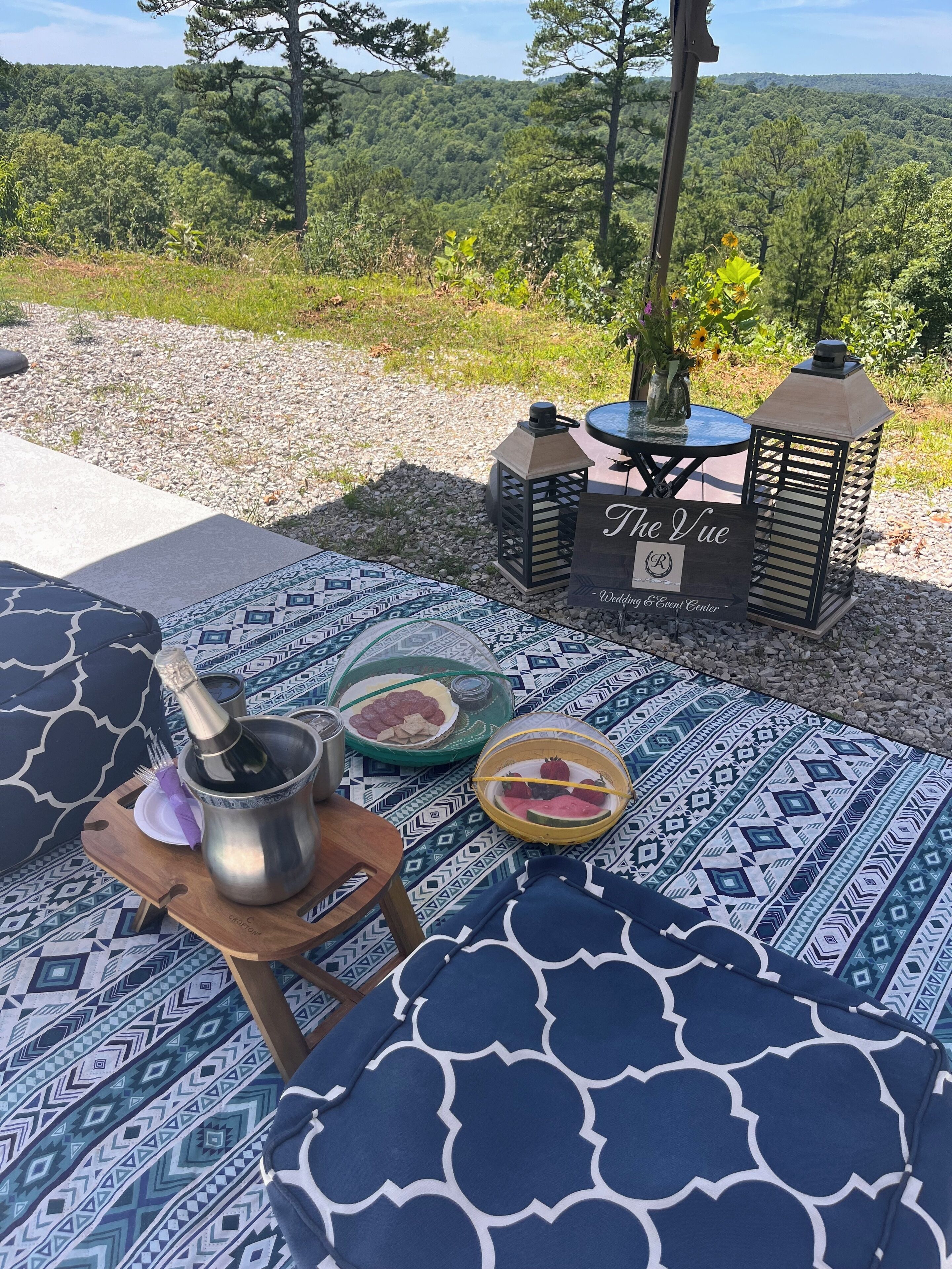Un espace extérieur aménagé avec des coussins à motifs, une table en bois sur laquelle sont posés une bouteille et des en-cas, et une enseigne indiquant « The Vue Wedding & Event Center », le tout sur fond d’une colline boisée.