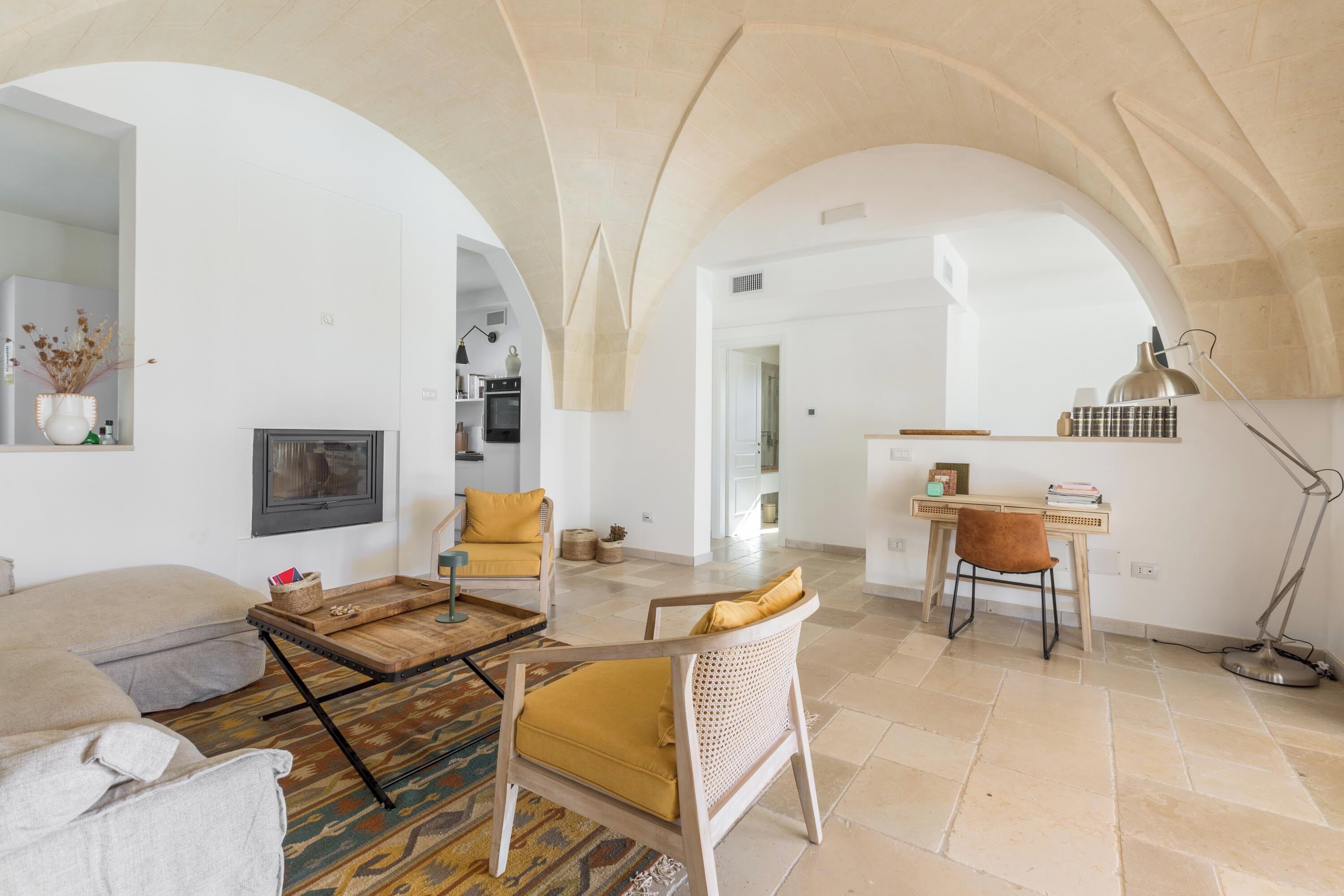 Un espacio habitable amplio con chimenea, una mesa de centro de madera y un sofá. Hay un escritorio con una silla y una lámpara. La habitación tiene un techo abovedado y vista a otra estancia.