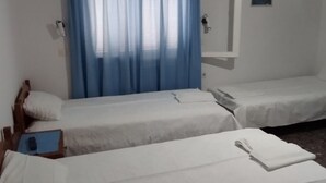 Una habitación con tres camas individuales, cada una con cabecera de madera, ropa de cama blanca y cortina azul. Una pequeña mesa con un libro y un control remoto está junto a una de las camas.
