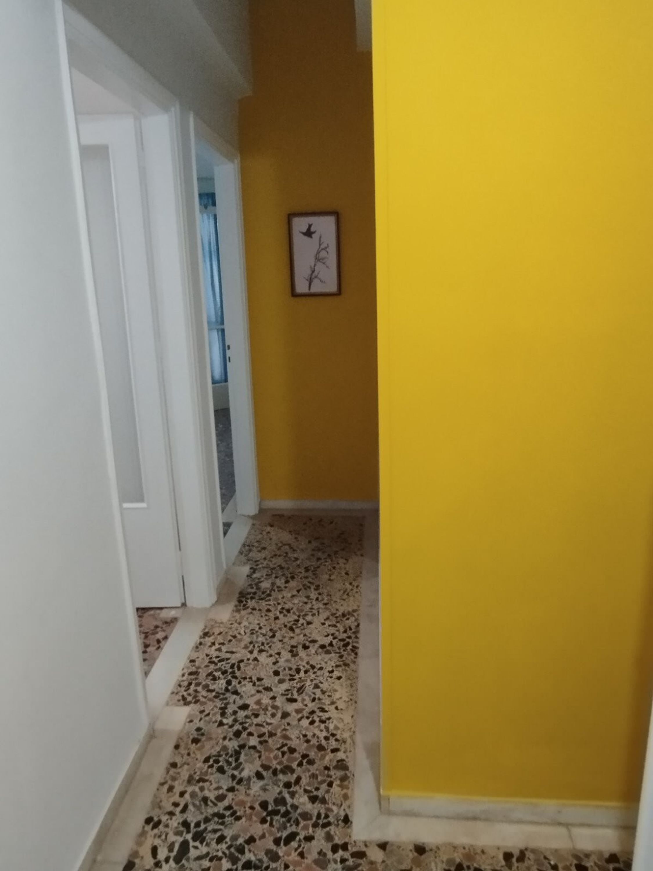 Un couloir avec un mur jaune et un tableau encadré représentant un oiseau et une branche.