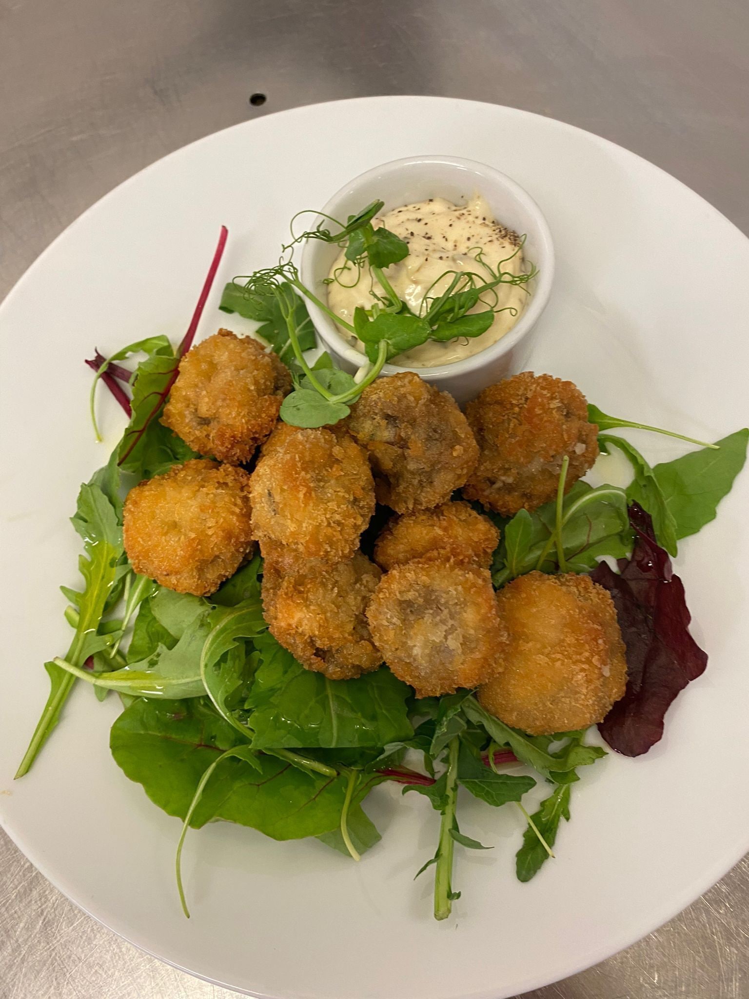 Un plato de alimentos empanizados y fritos, posiblemente croquetas, sobre una cama de hojas verdes mezcladas y acompañado de un recipiente pequeño con salsa para mojar.