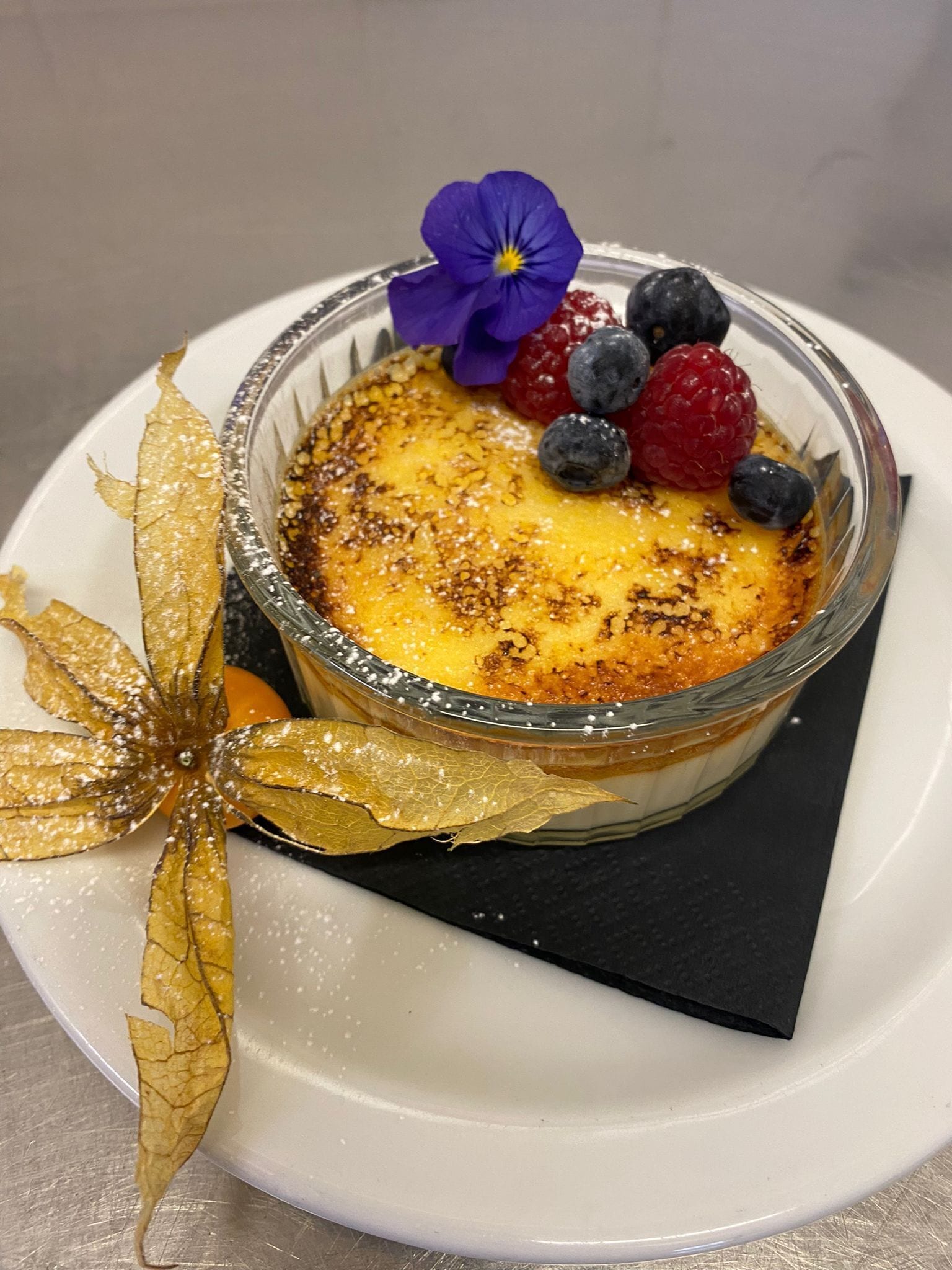 A dessert in a glass bowl with a golden crust, berries, and a purple flower on a white plate.