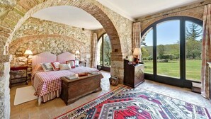 Une chambre avec des murs en pierre, un grand lit recouvert d’une couette à carreaux, une commode en bois et une vue sur l’extérieur grâce à de grandes fenêtres.