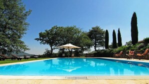 Une piscine entourée d’arbres et de grands cyprès, avec des chaises longues et un parasol.
