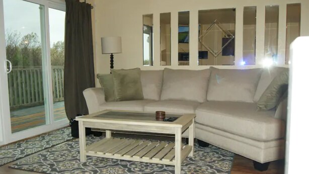 A living room with a beige sofa, a wooden coffee table, a patterned rug, and a sliding glass door leading to a balcony. - Soothing 2-bdrm condo near beach with WiFi and Fireplace (Ocean Shores)