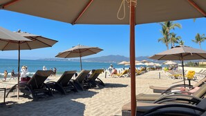 A beach with rows of sun loungers and umbrellas, a clear blue sky, and a view of the ocean and mountains in the distance. - Penthouse Luxury. WOW! Panoramic views of everything! BradAndJoeyRentals (Nuevo Vallarta)