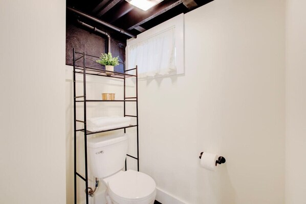 Un baño pequeño con inodoro blanco, una estantería metálica con planta en maceta y una toalla blanca colgada en la pared.