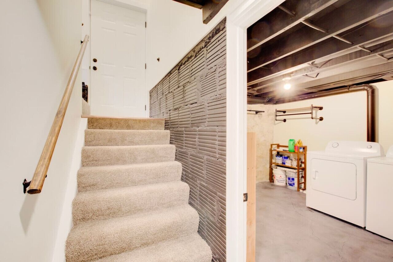 Un escalier menant à une buanderie avec une porte blanche, un mur en briques et un plafond où les conduits sont apparents.