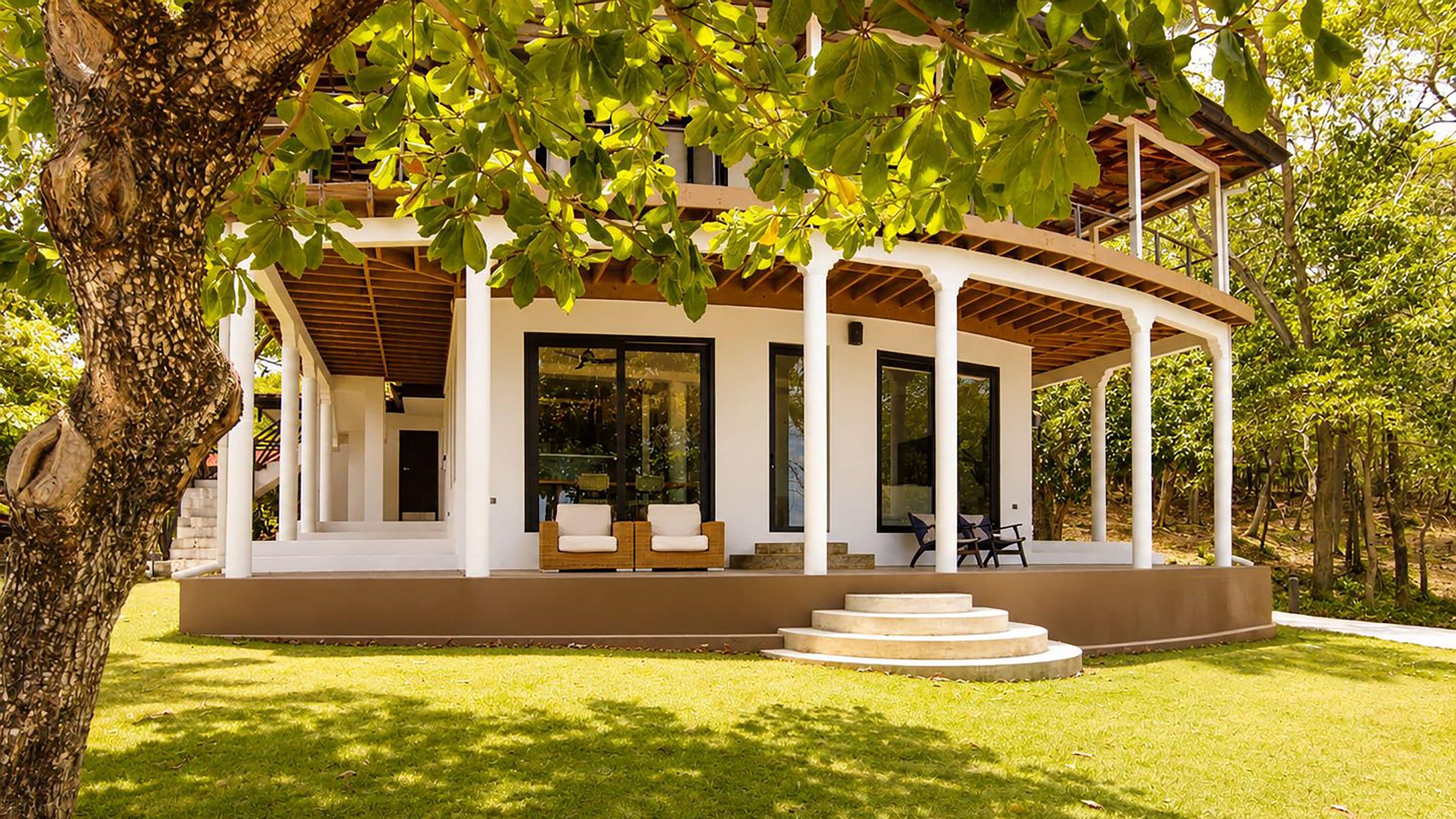 A covered outdoor seating area with a modern design, a staircase leading to a door, and a lush green lawn.