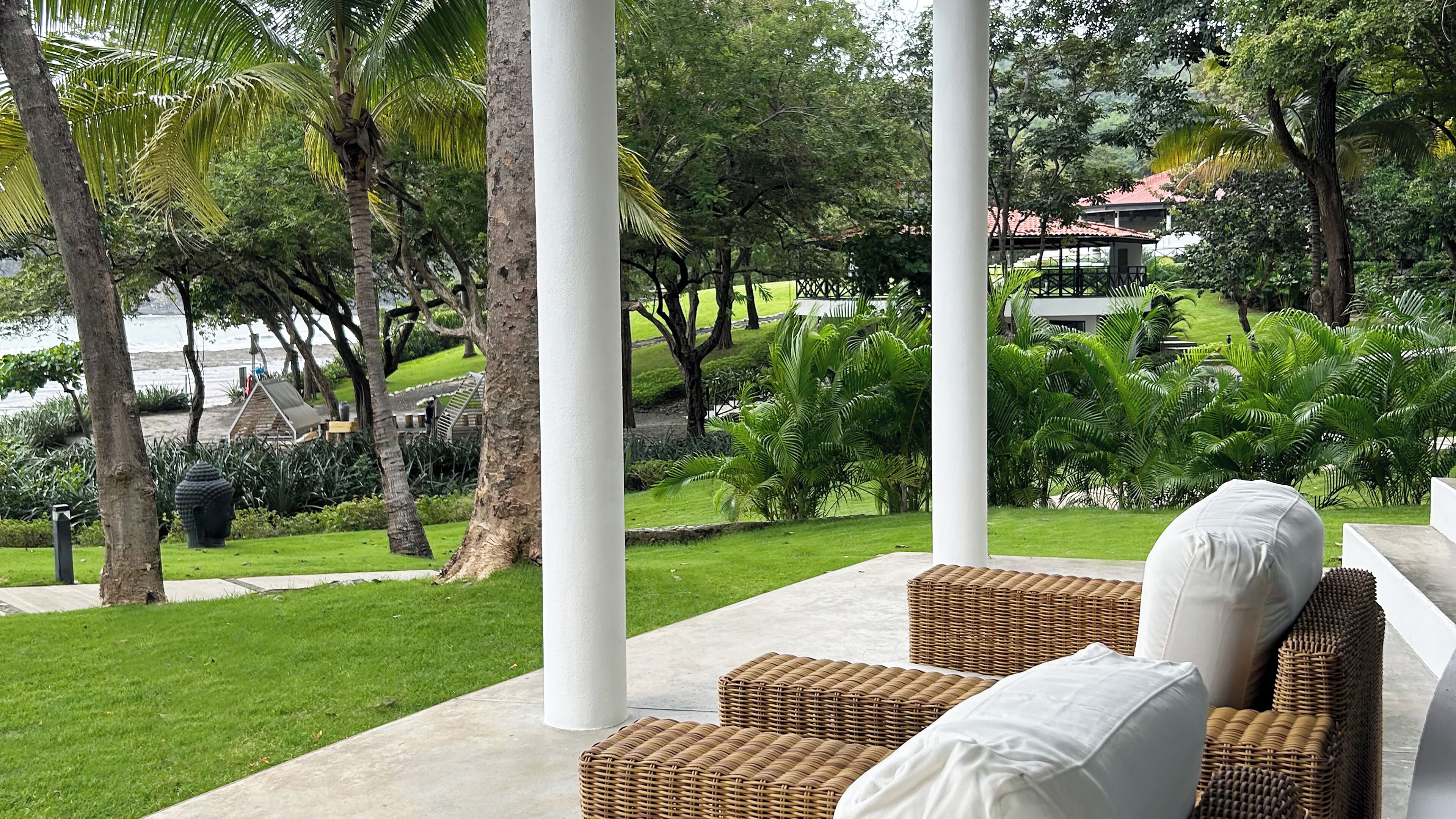 Un espace extérieur paisible, aménagé avec des meubles en osier et des coussins blancs, entouré d’une végétation luxuriante et de palmiers, et offrant une vue sur un jardin paysager et un plan d’eau.