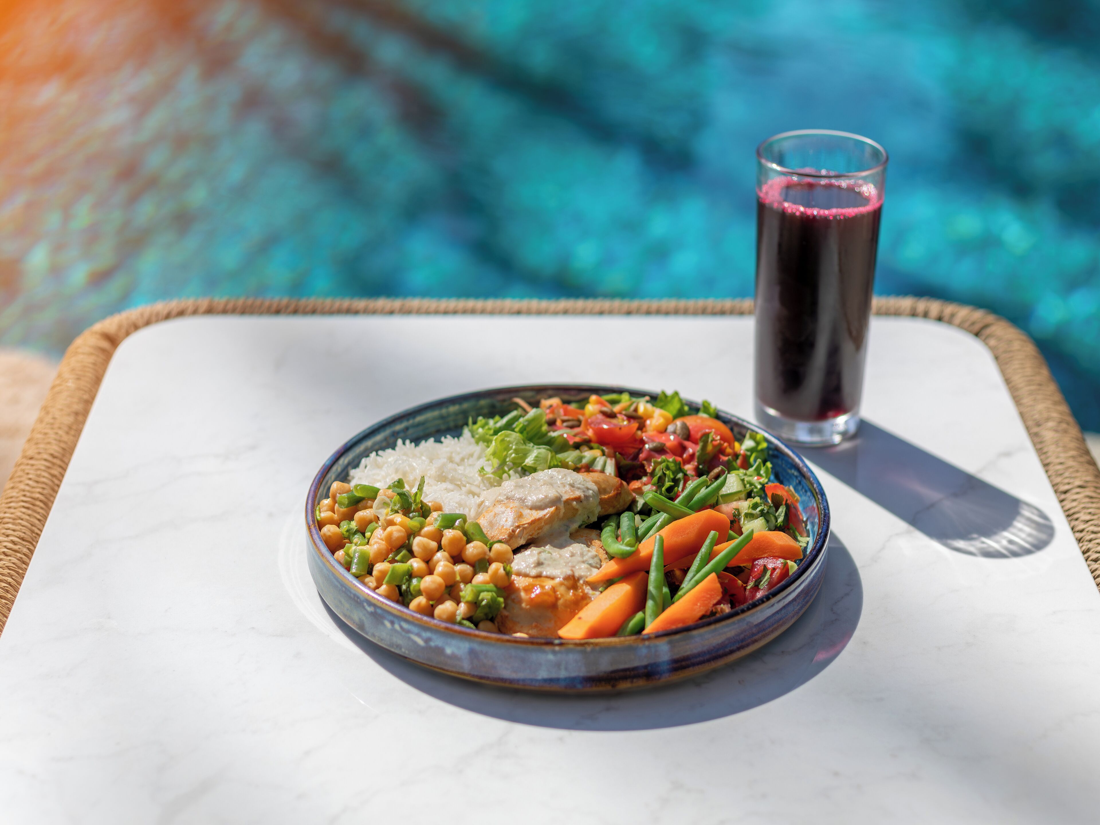 A meal with rice, vegetables, and a glass of red drink on a table by a pool.