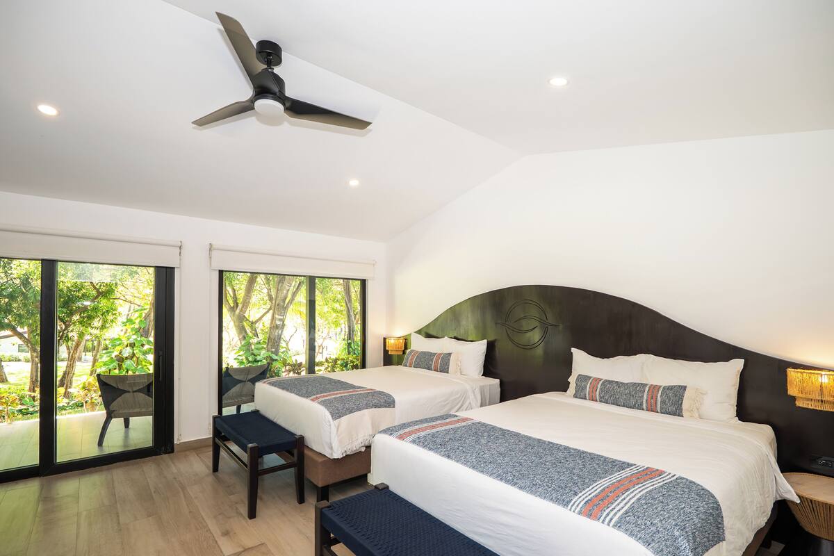 A hotel room with two beds, a ceiling fan, and large windows with a view of greenery.