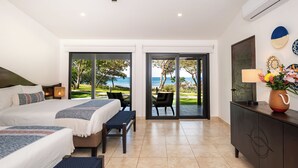 Une chambre d’hôtel comprenant un lit, deux tables de chevet, un bureau avec une chaise, un ventilateur de plafond et une vue sur la mer, visible à travers des portes vitrées.